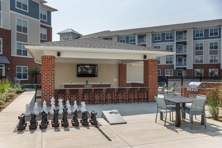 Attain at Towne Centre apartment homes with Outdoor patio area with seating, a bar, TV, grill, giant chess set, and apartment buildings in the background.