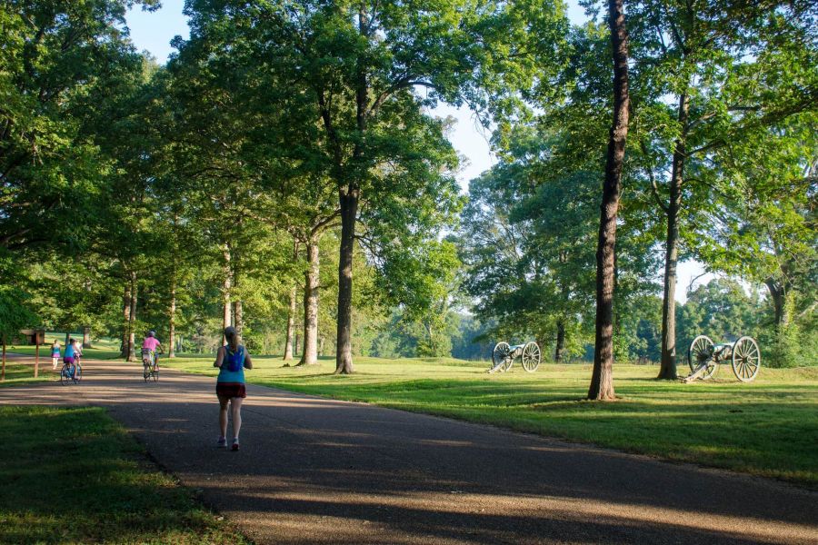 Attain at Towne Centre apartment homes with People walk and bike on a paved path in a sunny, tree-filled park with historic cannons on the lawn.