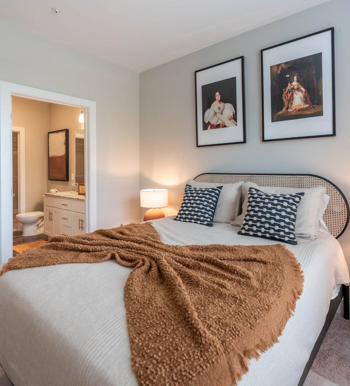 Attain at Towne Centre apartment homes with Cozy bedroom with a neatly made bed, two lamps, wall art, and an open door to a bathroom.