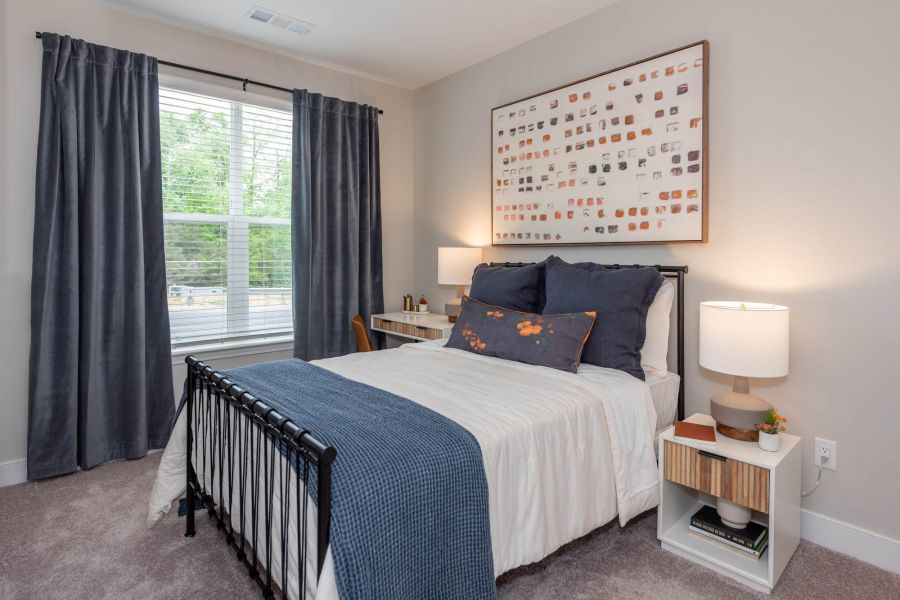 Attain at Towne Centre apartment homes with Bright bedroom with a black metal bed, blue bedding, white walls, large window, and abstract wall art.