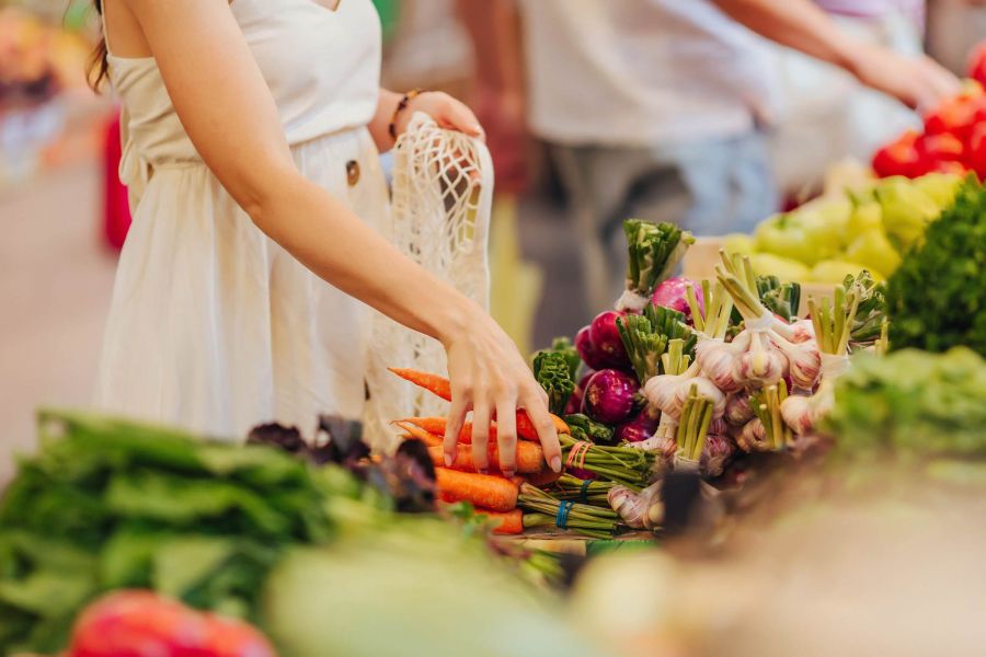 Attain at Towne Centre apartment homes with Woman in a white dress picking carrots at a market stall with various fresh vegetables.