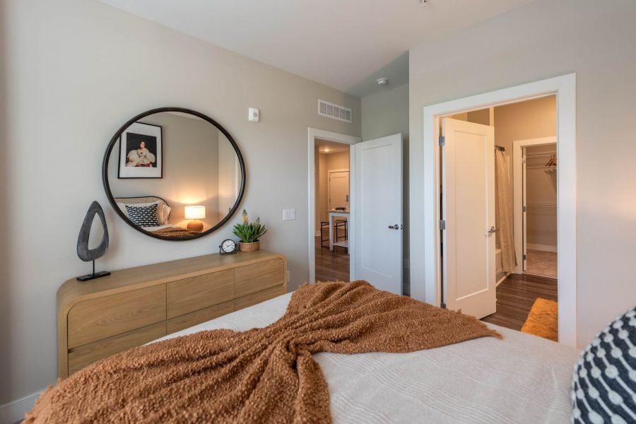 Attain at Towne Centre apartment homes with Modern bedroom with a wooden dresser, round mirror, and open door leading to a bathroom.