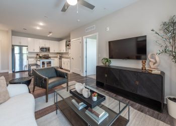 Attain at Towne Centre apartment homes with Modern living room with neutral tones, a TV on a black cabinet, armchairs, and an open kitchen in the background.
