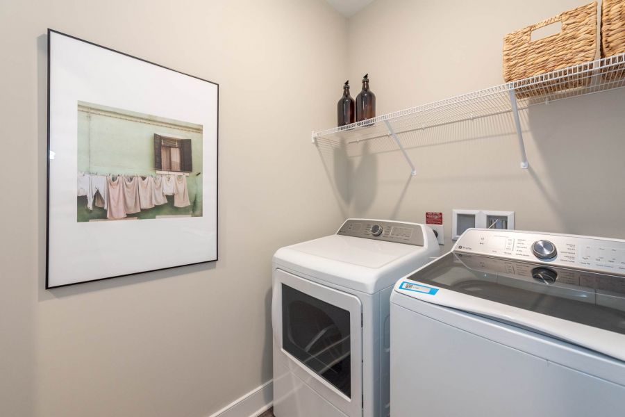 Attain at Towne Centre apartment homes with A laundry room with a washer, dryer, wall art, two brown bottles, and a wicker basket on a wire shelf.