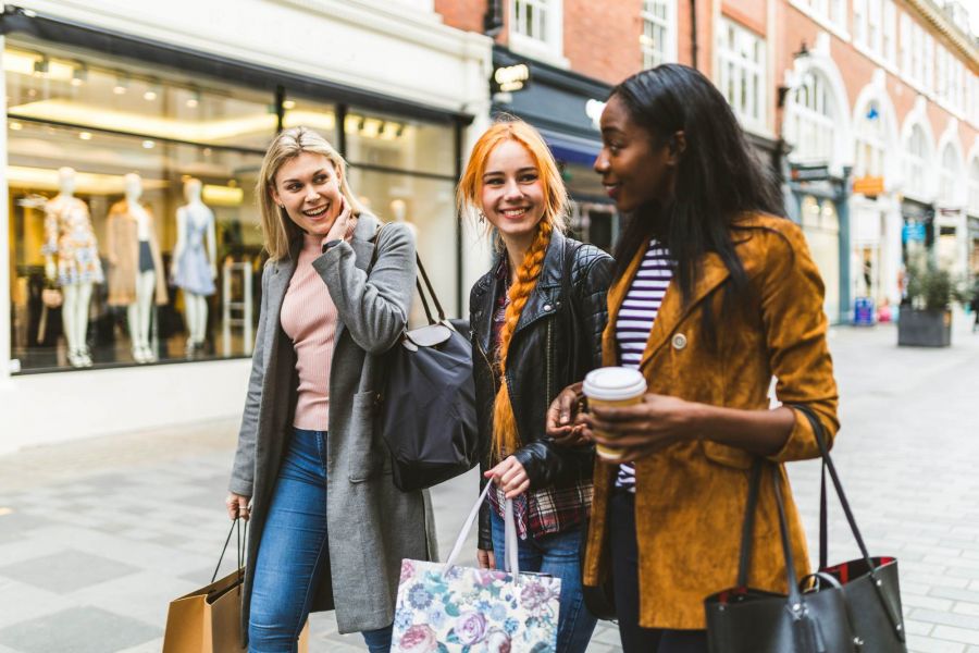 Attain at Towne Centre apartment homes with Three young women smile and walk with shopping bags on a city street in front of clothing store windows.