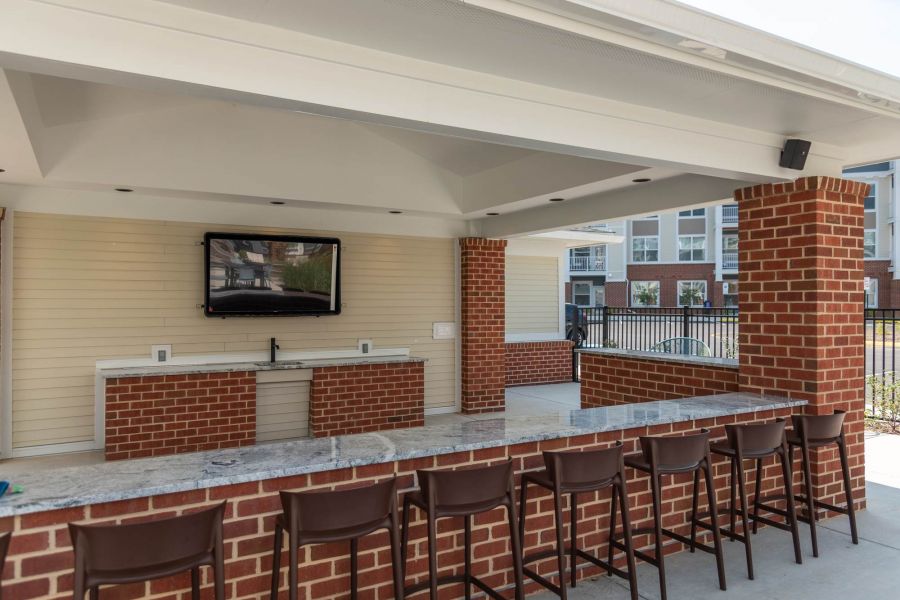 Attain at Towne Centre apartment homes with Outdoor bar area with brick and granite counter, brown chairs, wall-mounted TV, and nearby pool.